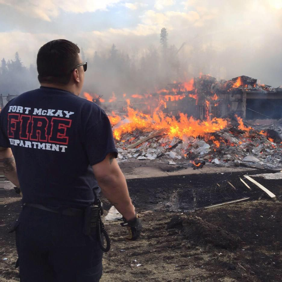 A man in a fire department-branded shirt stands with his bck to the camera looking at a fire and wreckage of a building