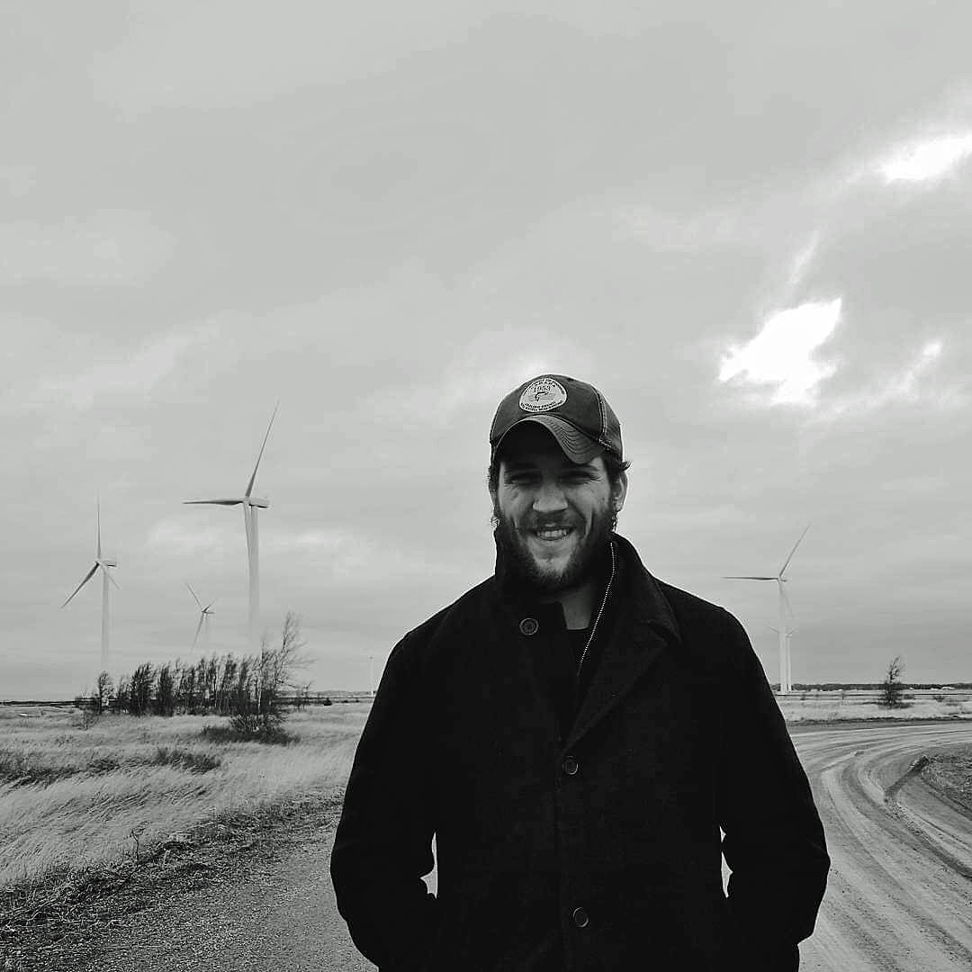 A man in a baseball cap and jacket stands in front of wind turbines