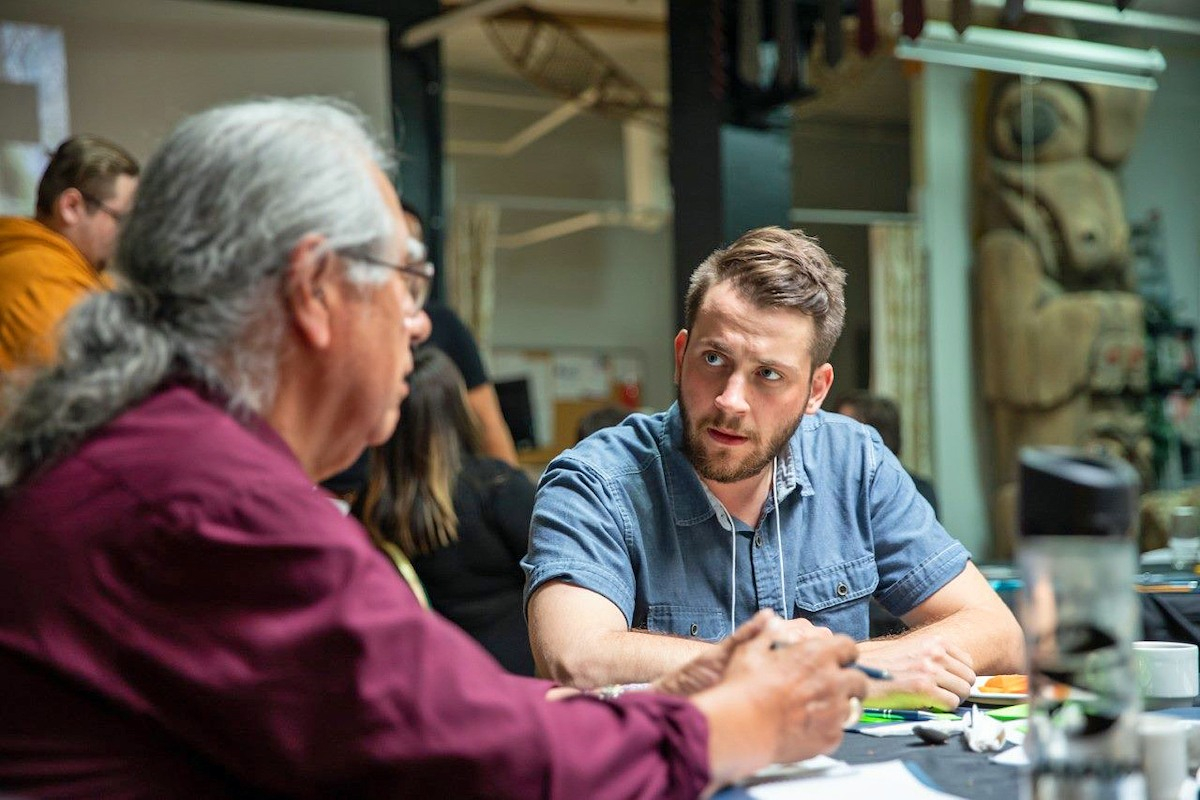 A blond man with facial hair sits at a table, listening to an older man with a ponytail to his right