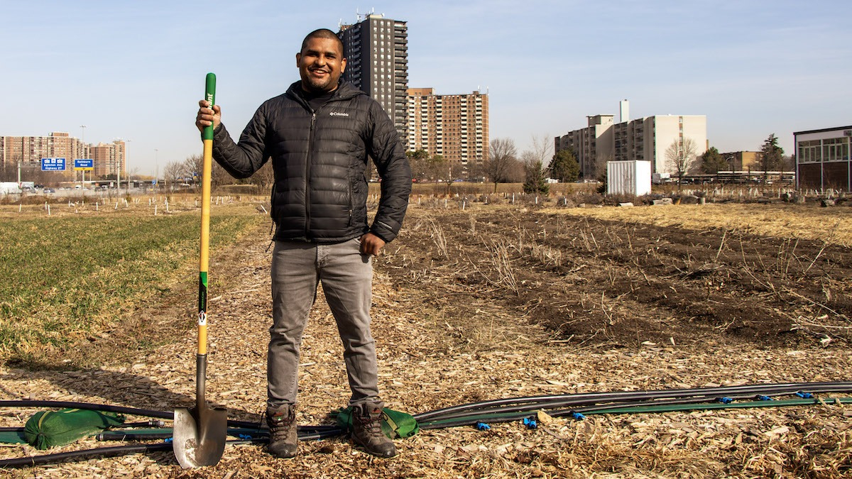 A man holding a shovel stands in front of an empty field with highrise buildings in the background