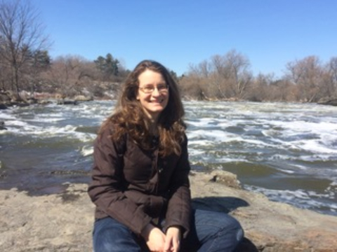 A woman with long hair sitting outdoors on rocks with a river behind her. 