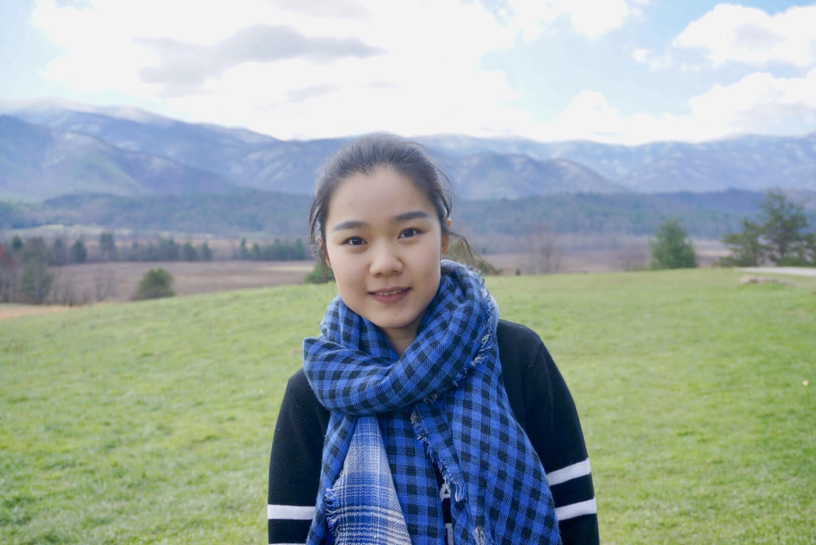 A woman wearing a blue checkered scarf stands, smiling in front of a backdrop of green hills and mountains.