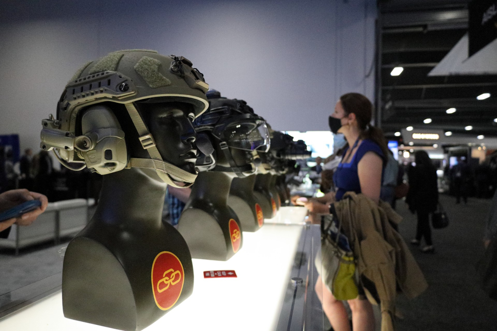 A line of military helmets sit ina  row, with a woman browsing in the background