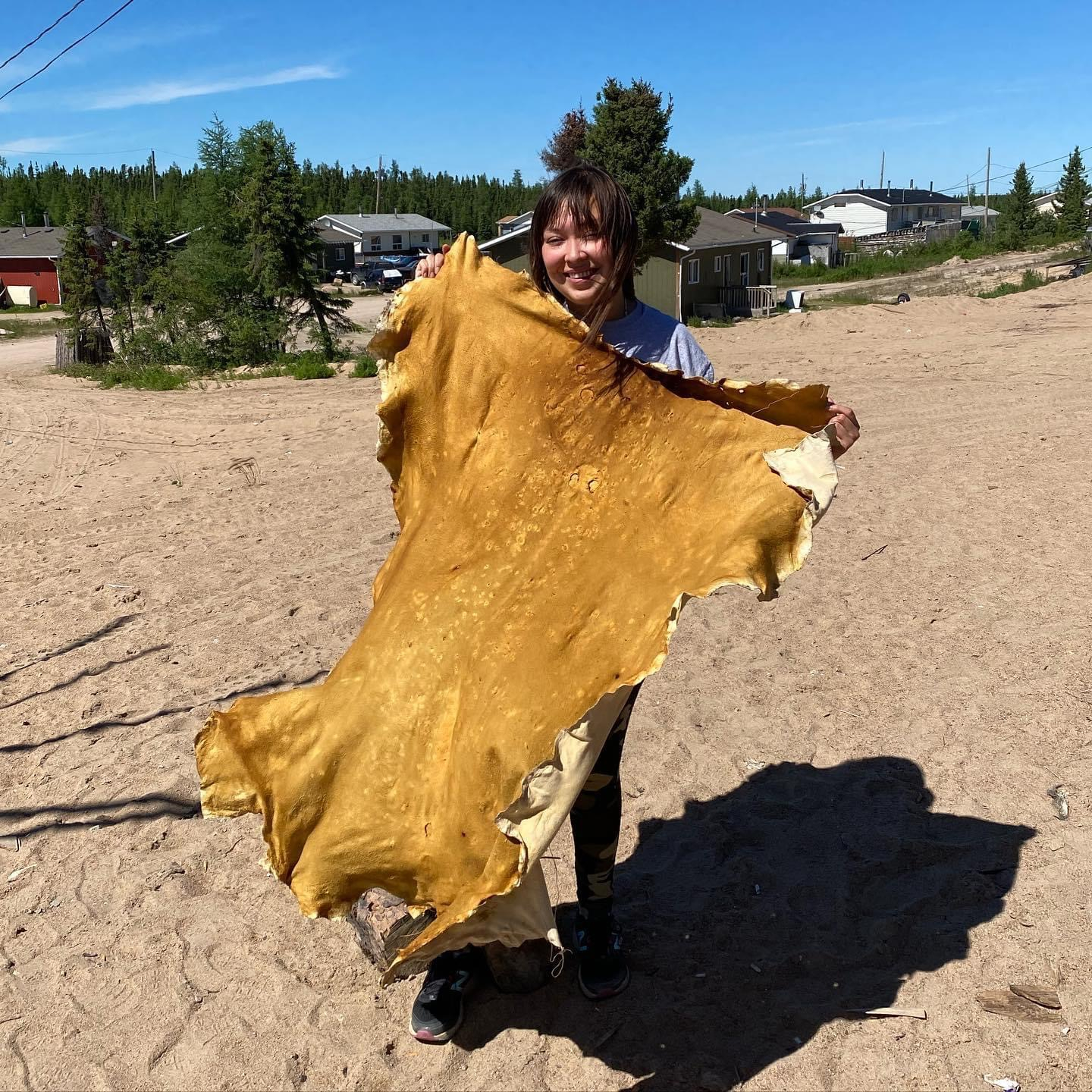 A woman stands with a big smile, holding up a deer hide she worked with using traditoinal methods