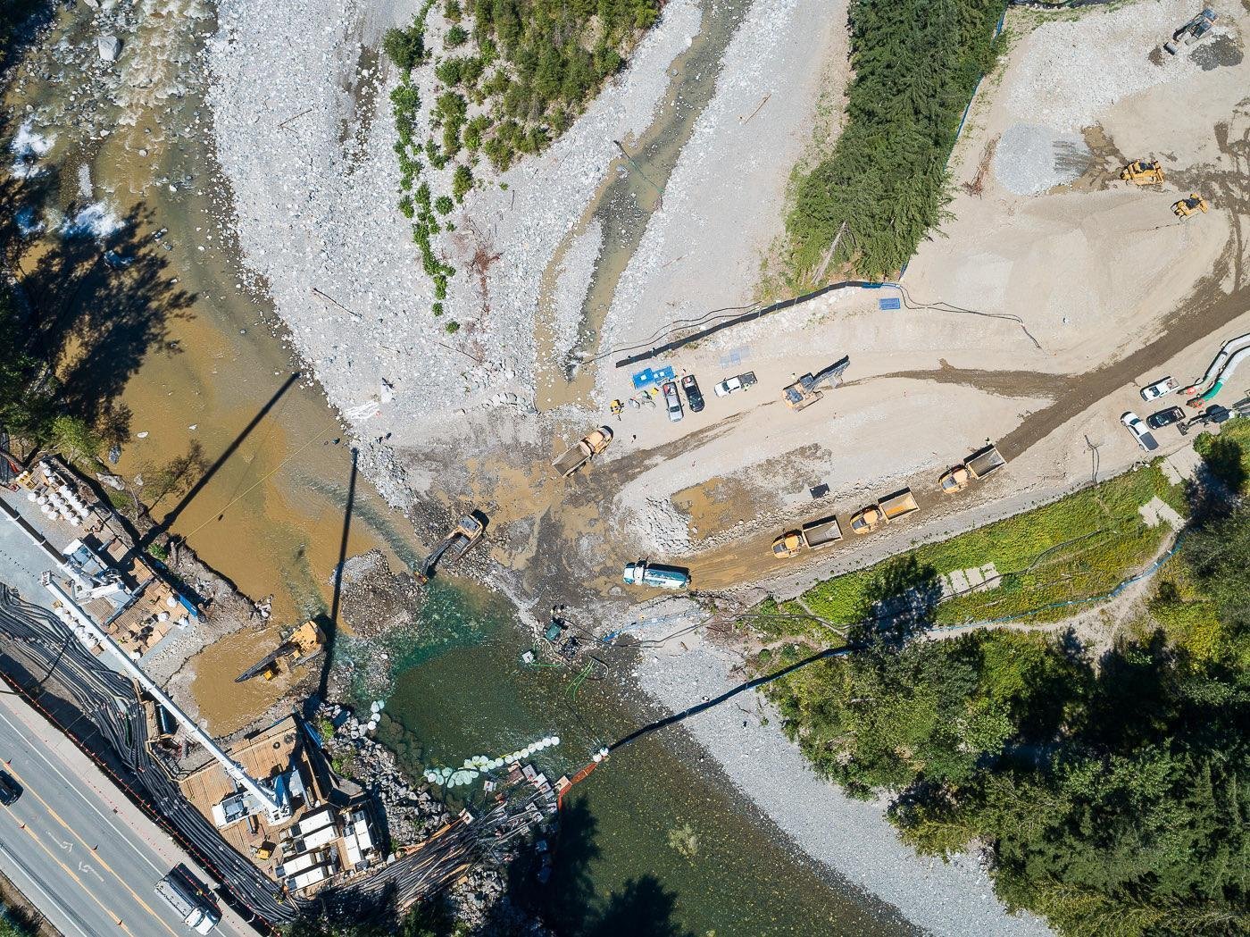 A birdseye view of a Trans Mountain worksite in Hope, B.C.. Excavators can be seen digging in the water, and downstream, in the upper left corner of the image the water is very muddy and turbulent due to pumps transporting water downstream.