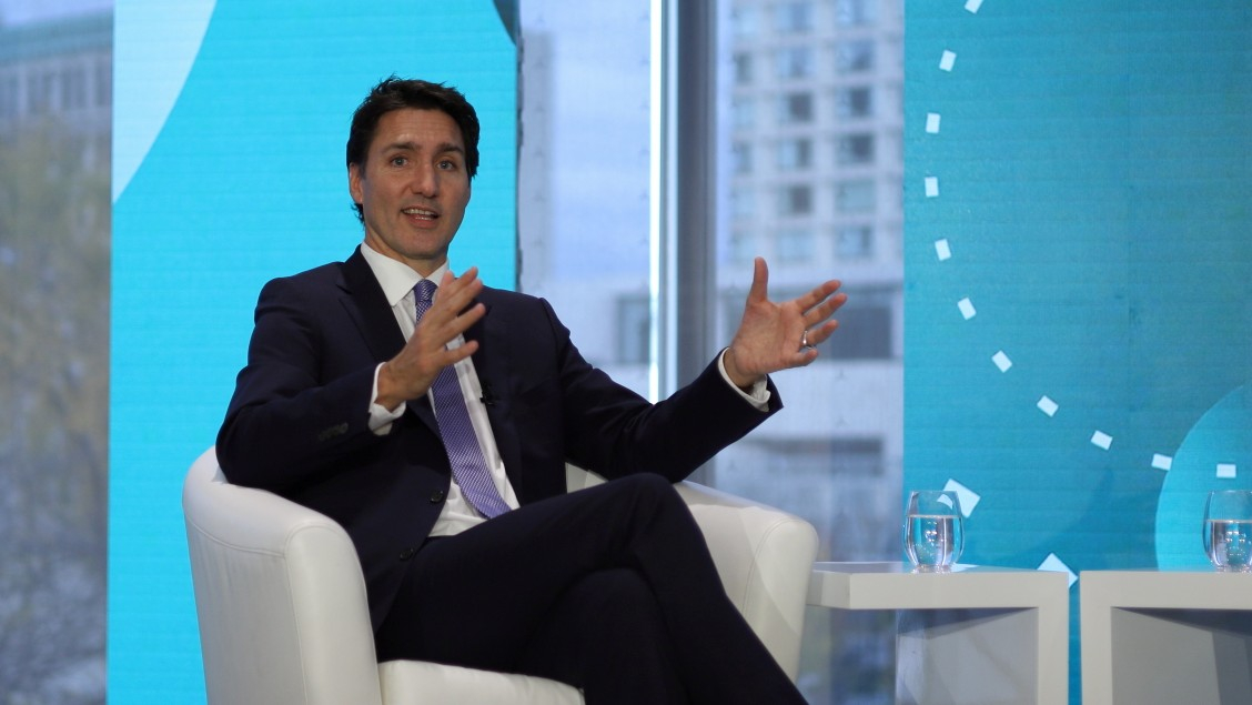 The prime minister of canada (a white man) sits in a white armchair, shown from the shin up, gesturing with both hands