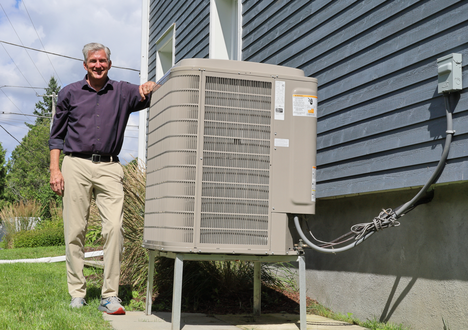 A man standing with his arm on an air source heat pump outside his blue house