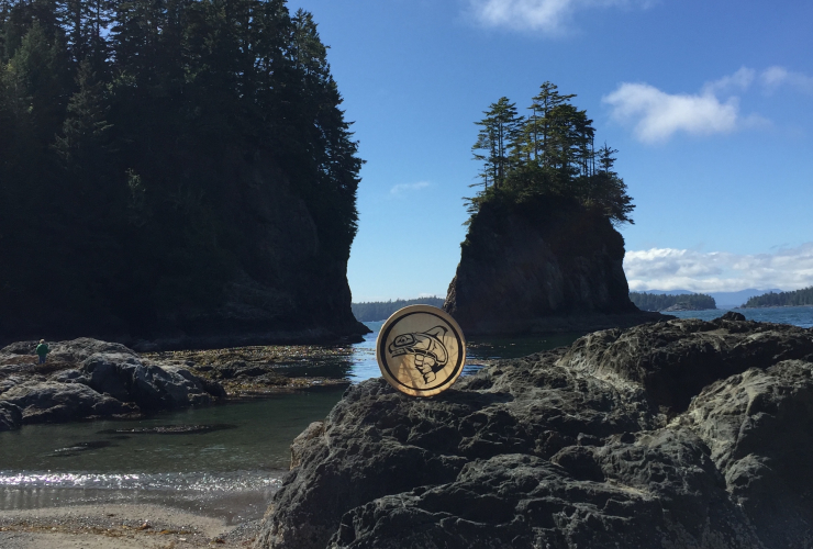 This is the setting of the ancient Huu-ay-aht village of Kiixin on Vancouver Island, with a drum featuring the Huu-ay-aht logo. 