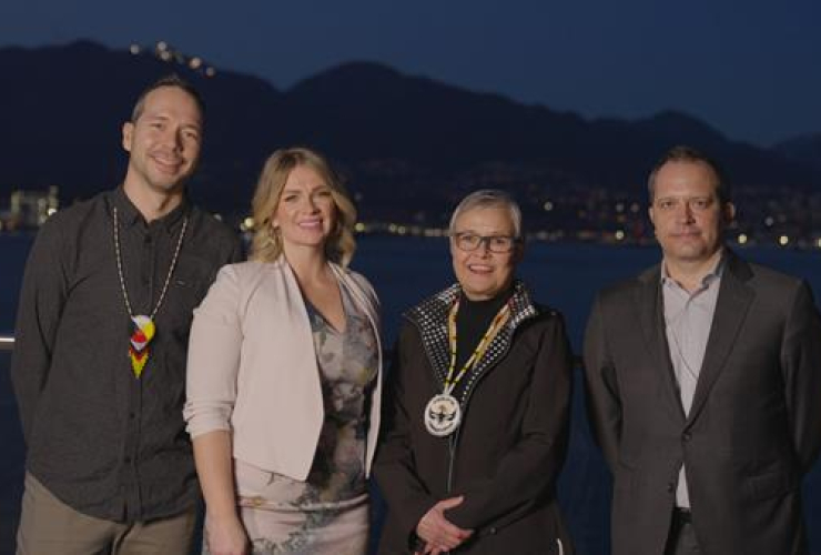Four people stand side by side in Vancouver with a dark blue background of ocean and mountains in the evening.