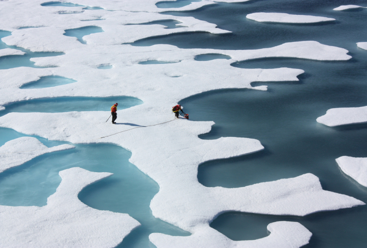 Two people dressed in high visibility colours stand out brightly on a vast expanse of melting ice with large pools of water dispersed throughout. They appear to be taking measurements or samples of the ice. 