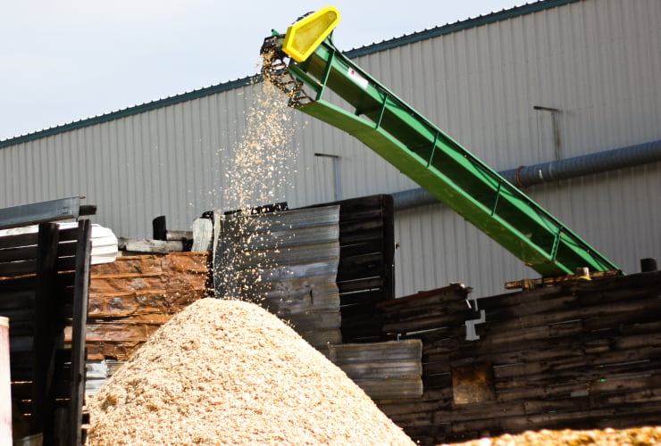 Tall green piece of machinery sits at a diagonal with wood shavings coming out the top and landing in a pile below.