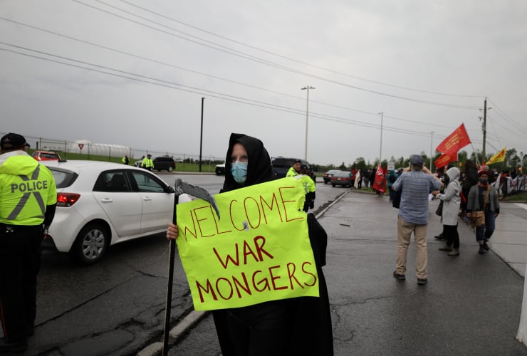 A person dressed as the grim reaper holds a sign that says "Welcome war mongers"