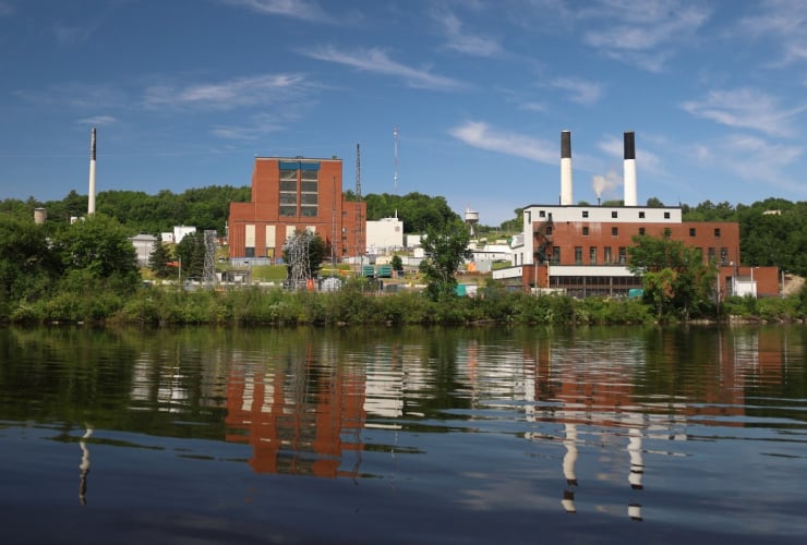 Brick buyildings and smoke stacks of the Chalk River nuclear laboratory are visible from the river on a bright, sunny day