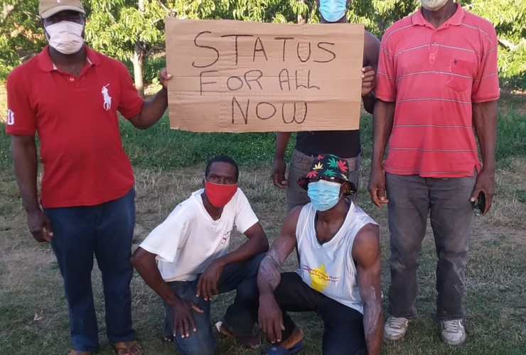 Three migrant workers stand holding a carboard sign that says "status for all now" while two more workers crouch below the sign
