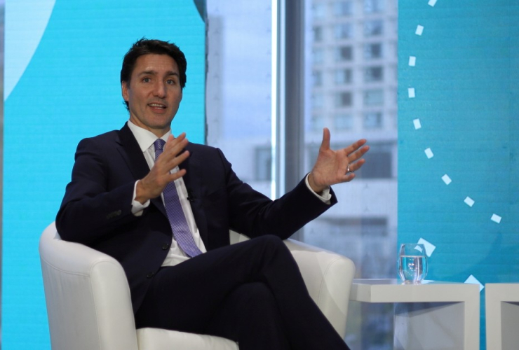The prime minister of canada (a white man) sits in a white armchair, shown from the shin up, gesturing with both hands
