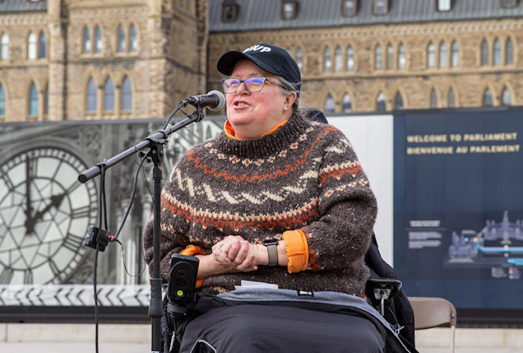 A woman in a power wheelchair speaks into a microphone outside Parliament buildings in Ottawa, Ontario.