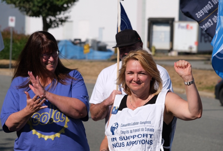photograph of three people, one woman with short blonde hair and a white shirt that says "in support" holds her fist up