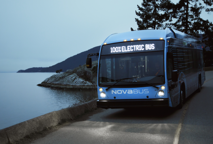 a blue bus that says "100% electric" driving on a road beside a body of water at dusk