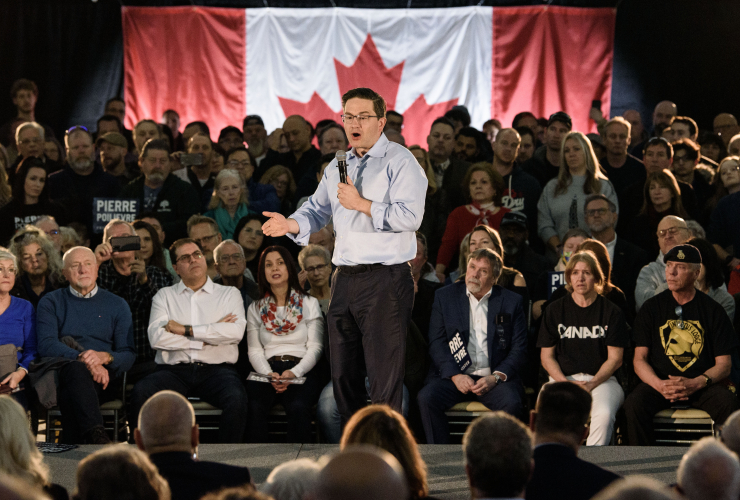 Conservative party leader Pierre Poilievre stands on a platform at a rally, with supporters standing all around him