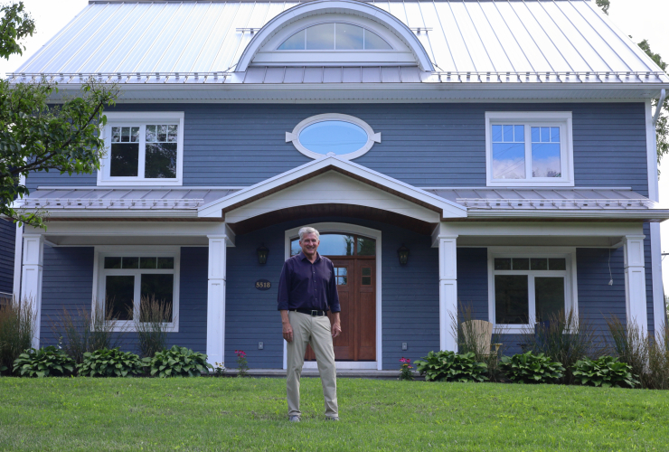 A man in a long sleeve collared shirt stands in front of a dark blue house on a bright, August afternoon