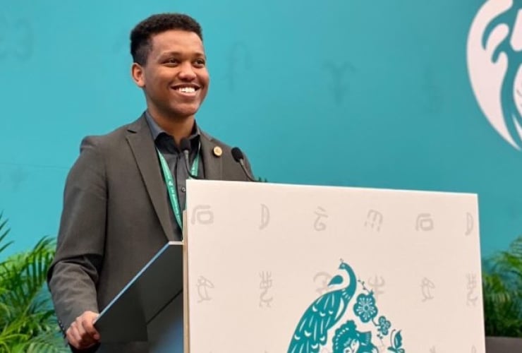 A young smiling man stands at a podium in front of  a turquoise backdrop